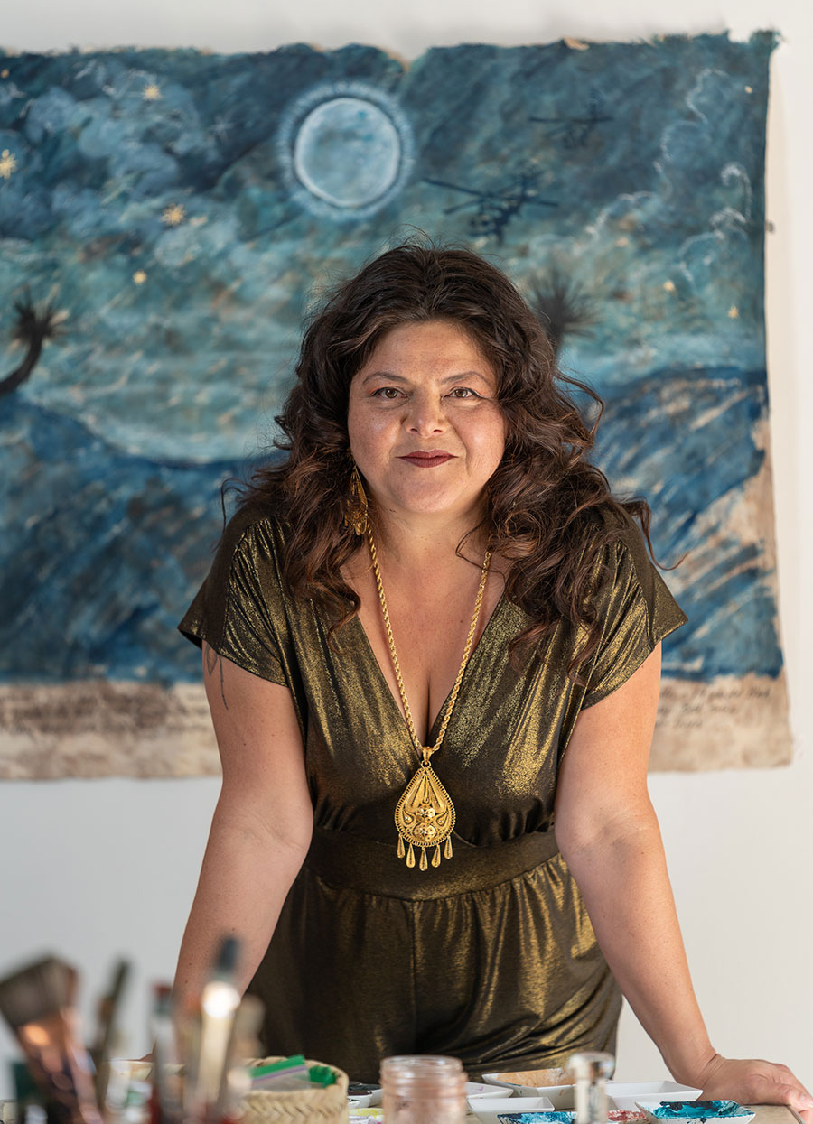 Sandy Rodriguez stands with painting materials in the foreground and a blue watercolor behind her on the wall.