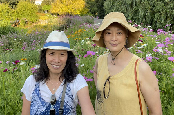 Two people smile at the camera in front of a field of pink and white flowers.