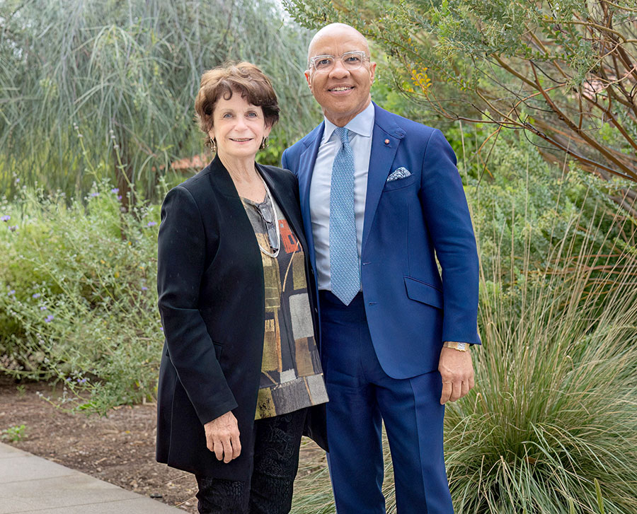 Two people pose for the camera in front of a garden of green brush and trees.