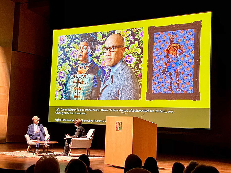 An audience watches two people have a conversation on a stage, where a large graphic shows two paintings.