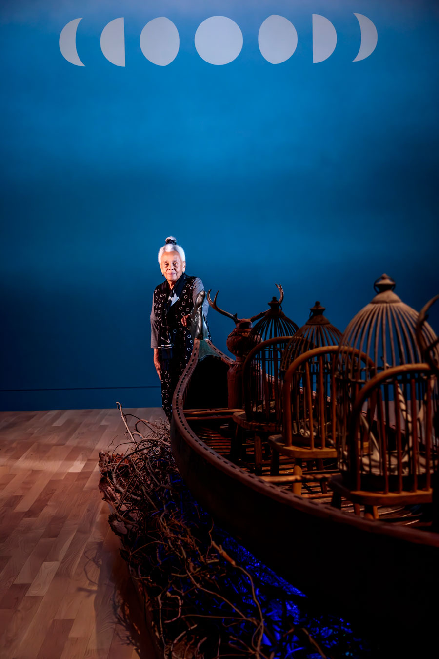 Artist Betye Saar stands near a wood canoe in a blue room with phases of the moon painted on the wall.