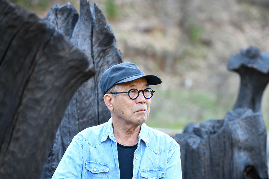 A man in a blue shirt and hat looks off camera, in front of a rock or tree formation.