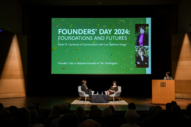 Two people sit in chairs on a stage in front of a large screen.