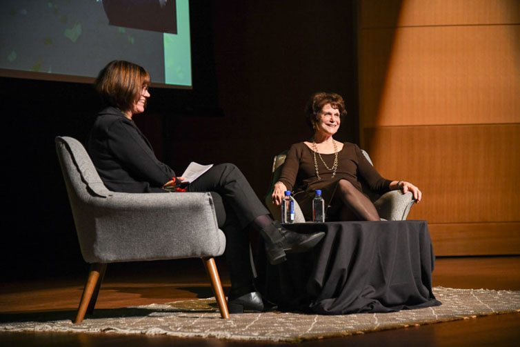 Two people sit in chairs on a stage.