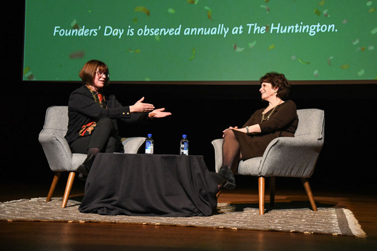 Two people sit in chairs on a stage in front of a large screen.