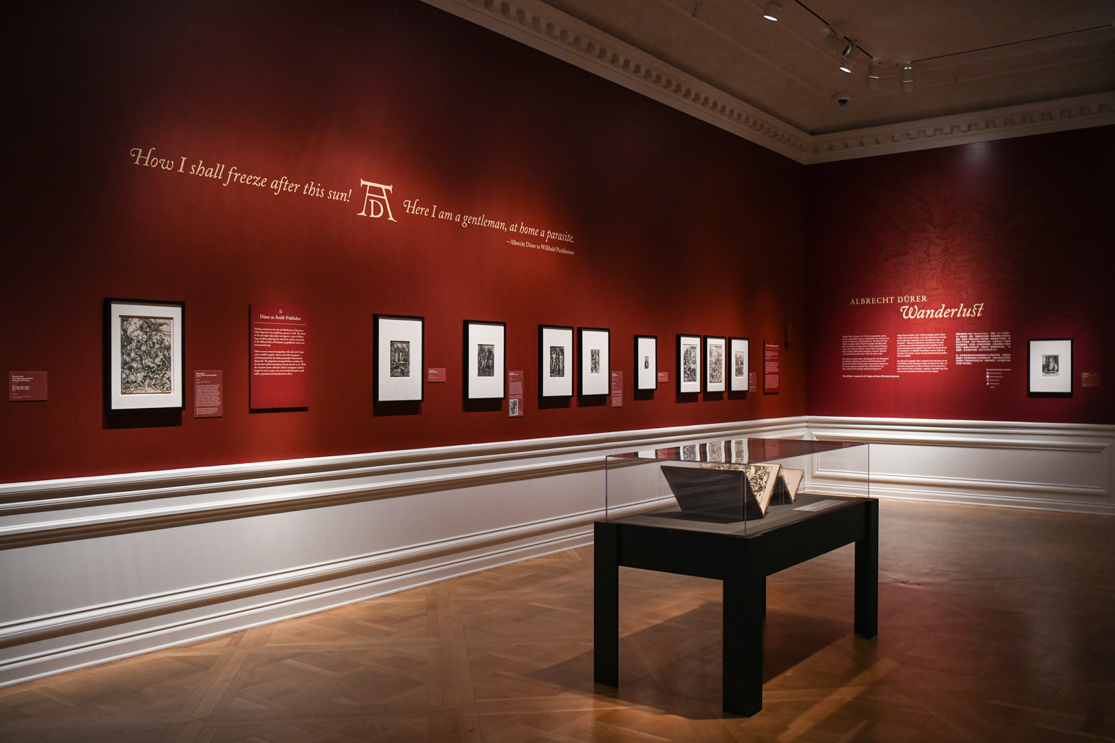 A gallery with dark red walls and beige text features matted artwork in black frames and a display case with two open books.