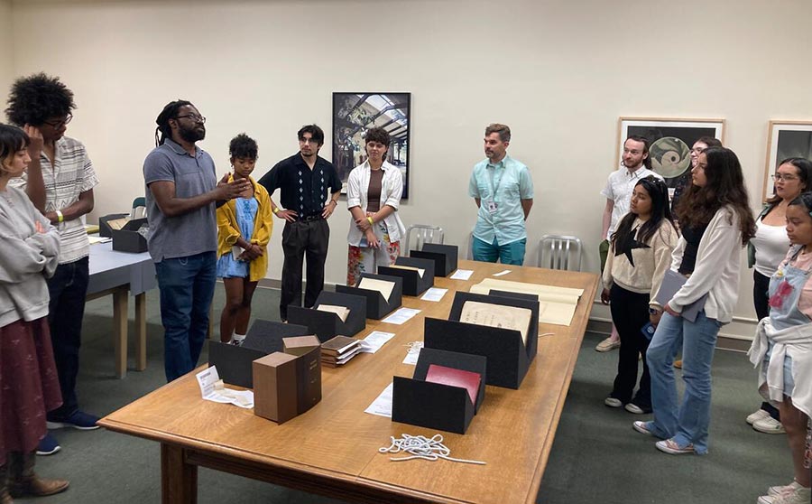 A group of people listen to a person speaking about books on a table.