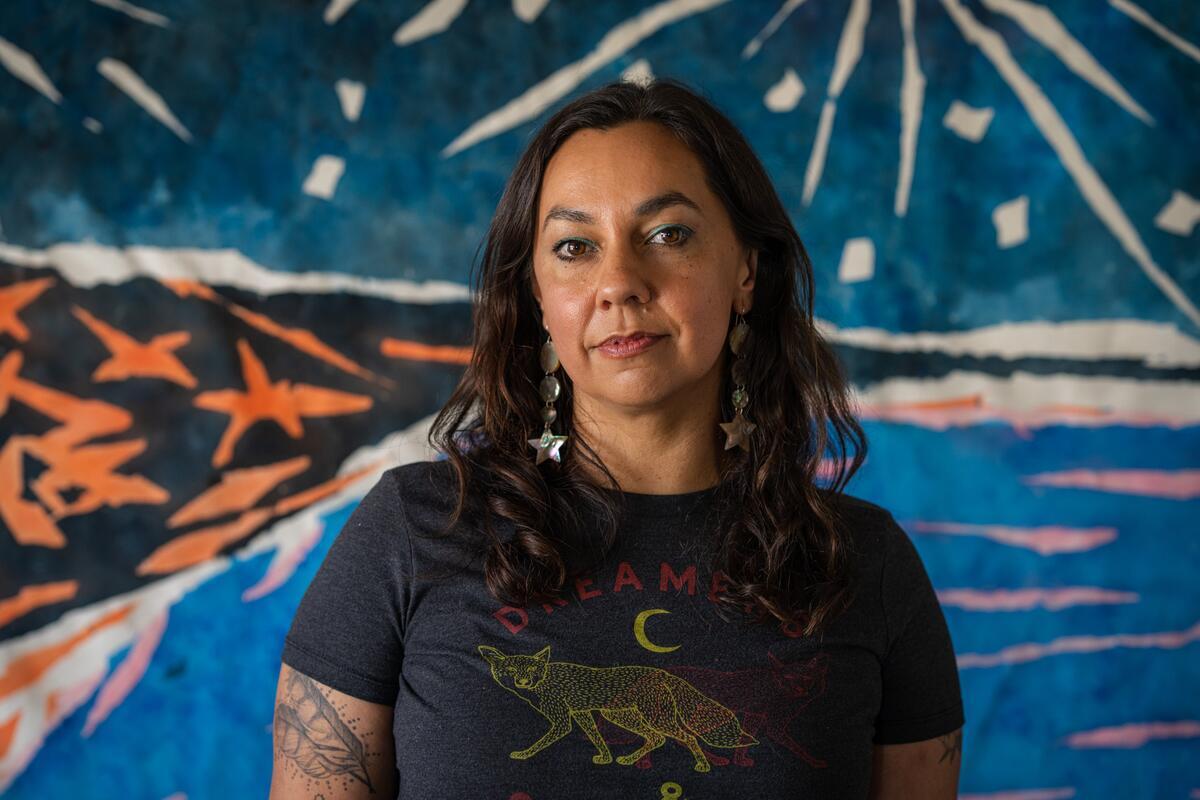 A person with wavy, brown shoulder-length hair stands in front of an abstract painting of a landscape with blue details.