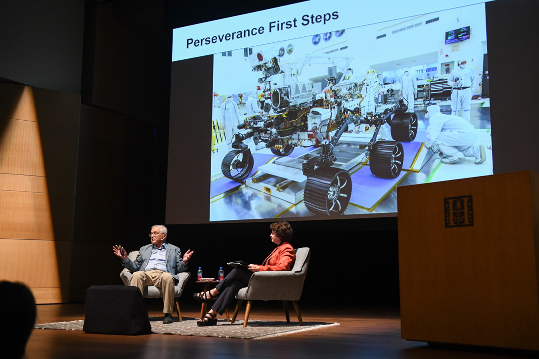 Two people sit on a stage in front of an audience, with a screen displaying an image of a robotic vehicle.