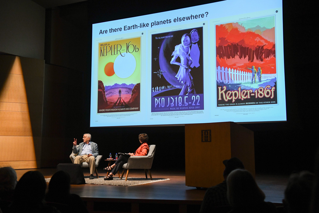 Two people sit on a stage in front of an audience, with a screen displaying three colorful posters depicting planetary travel.