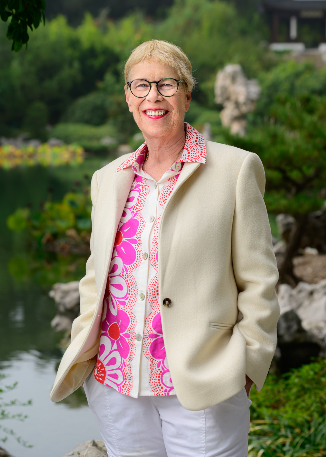 A smiling person in a pink floral shirt stands near water in a green garden. 