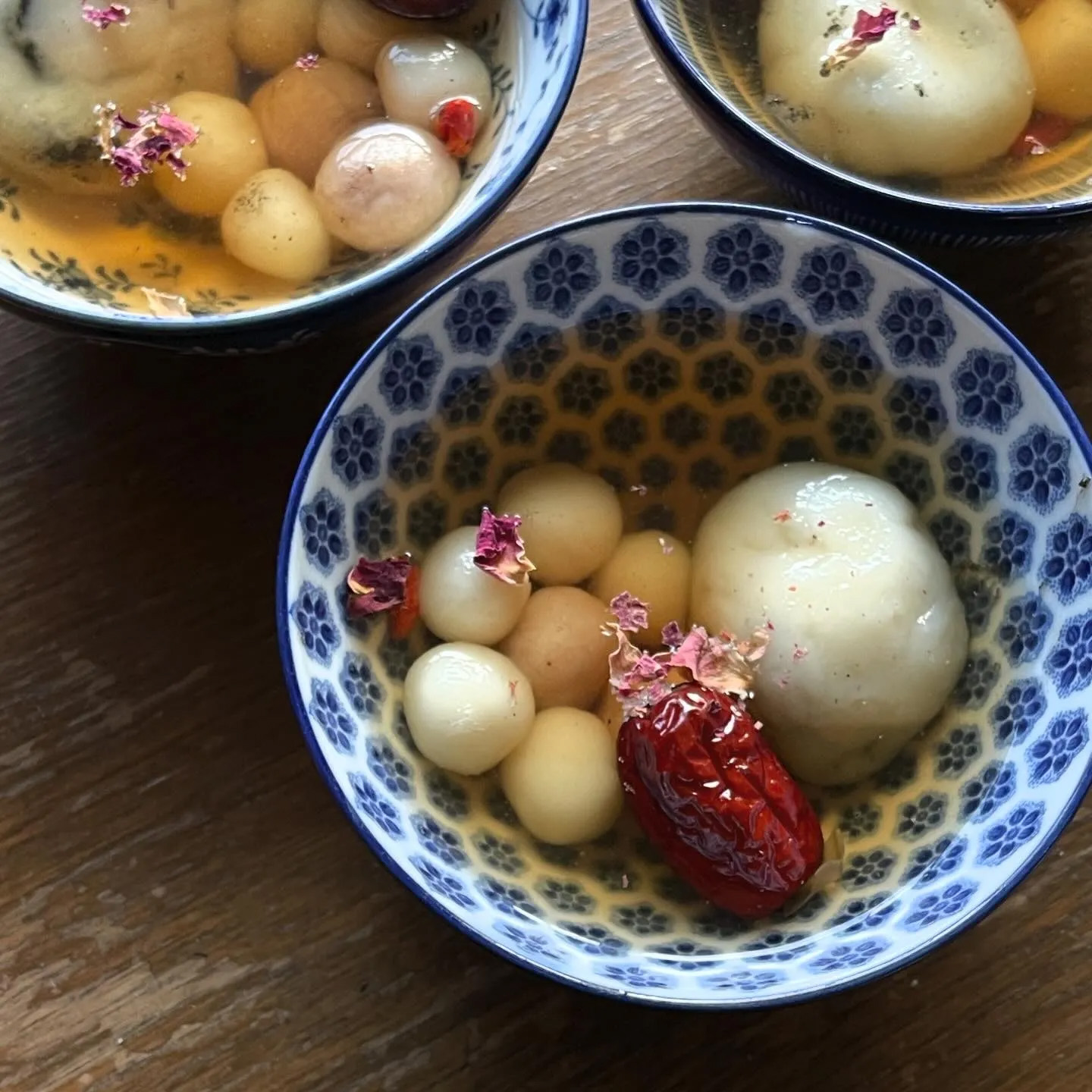 A blue and white ceramic bowl with clear-brown broth, white rice balls, red jujube, and flower petal garnish.