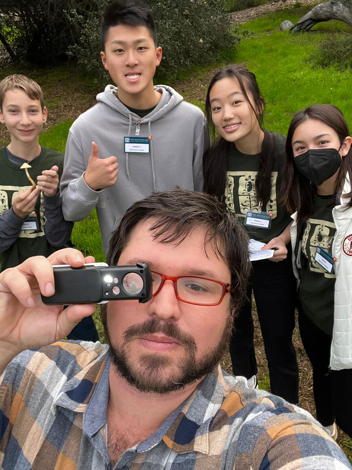 A person takes a selfie while holding a magnifying lens, with four people in the background.