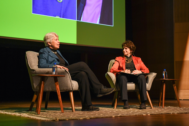 Two people sit on chairs on a stage in front of a green screen.