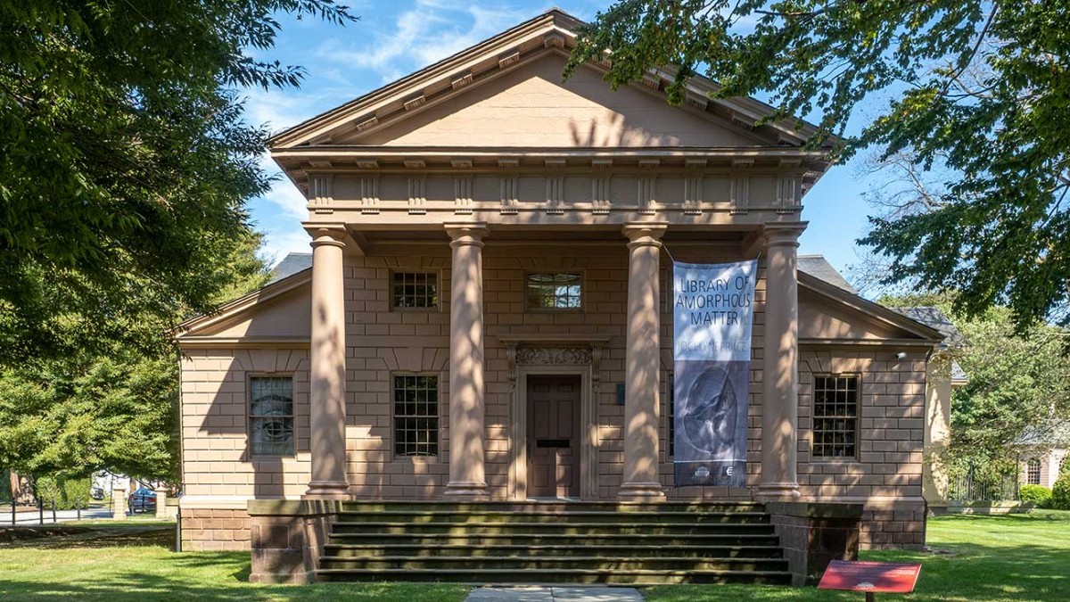 A tan-colored stone library with four large columns, surrounded by a green lawn and large trees.