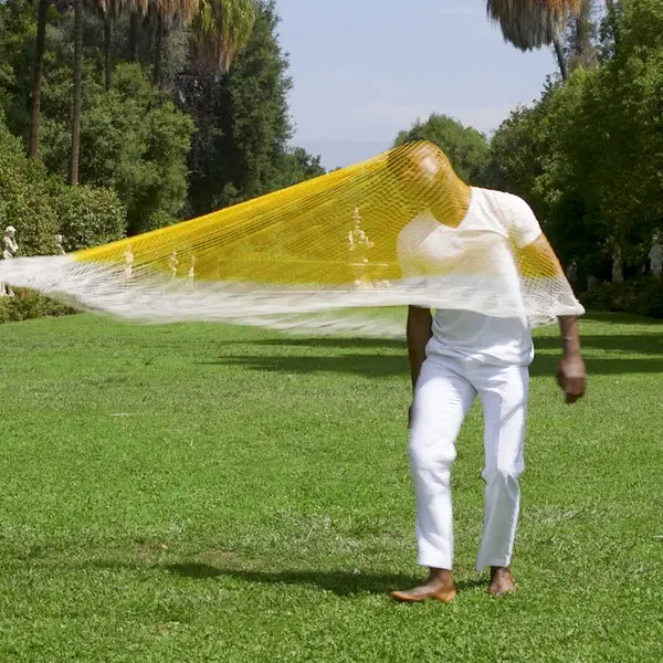 A Black person in white clothing spins a yellow net on their head and shoulders. The person is barefoot on a long, statue-lined lawn.