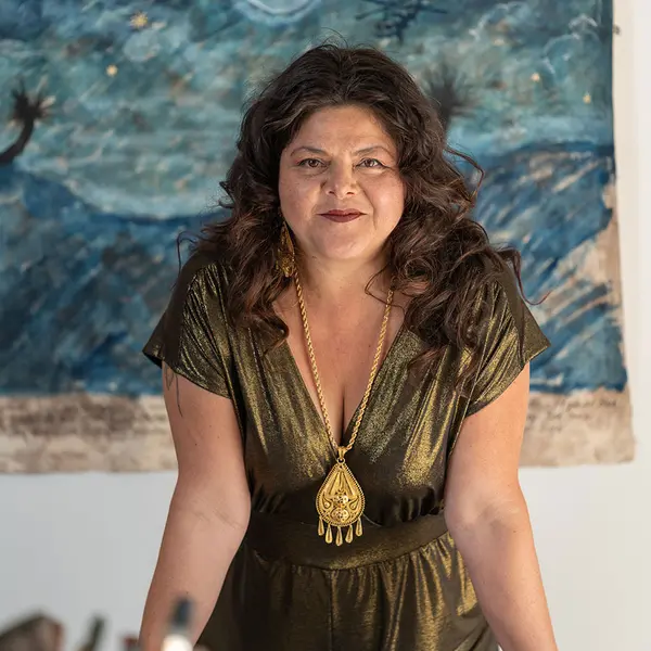 Sandy Rodriguez stands with painting materials in the foreground and a blue watercolor behind her on the wall.