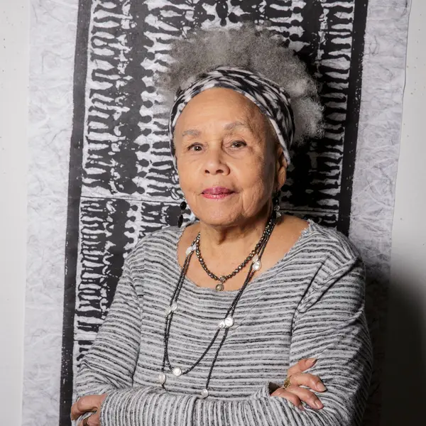 Artist Betye Saar, stands facing the camera with arms crossed in front of a pattern of bodies in silhouette.