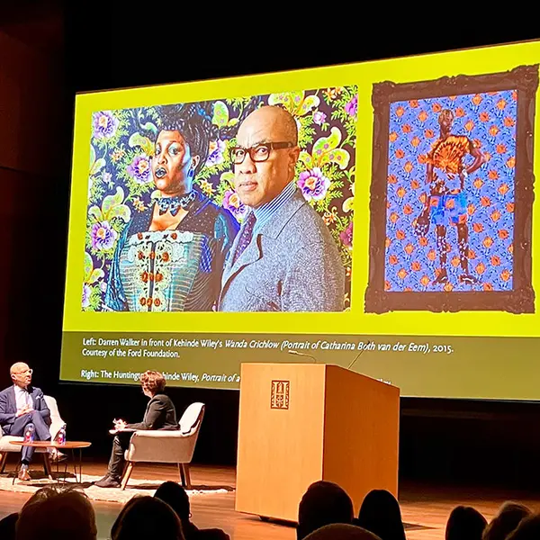 An audience watches two people have a conversation on a stage, where a large graphic shows two paintings.