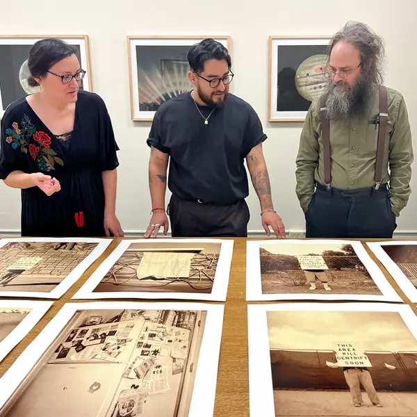 Three people stand over a table observing a series of photo prints.