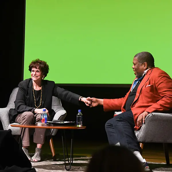 Two people on a stage, sitting next to each other and shaking hands.