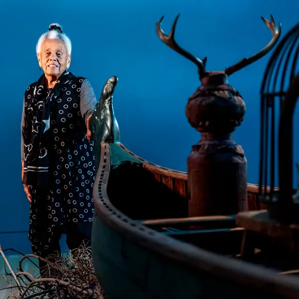 Artist Betye Saar stands near a wood canoe in a blue room.