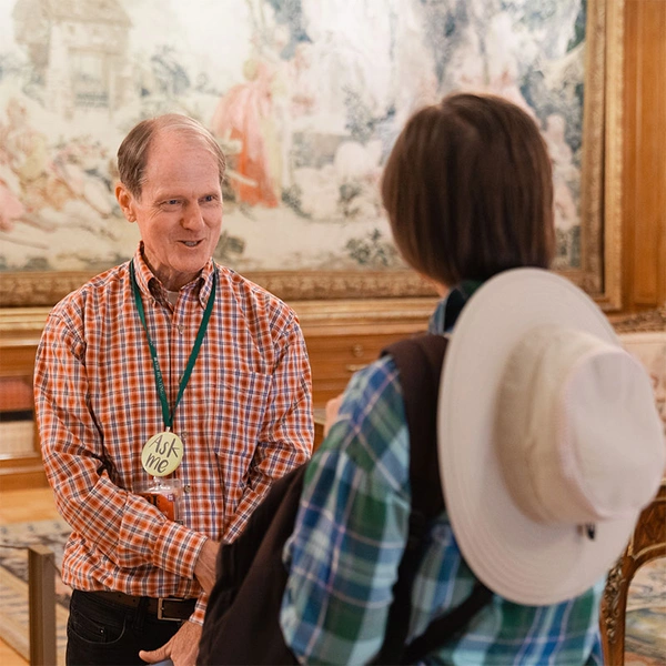 A person wearing an "Ask Me" badge talks to another person in front of a large hanging tapestry.