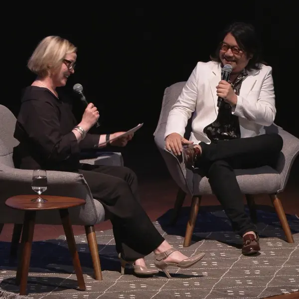 Two people holding microphones sit in chairs on a stage.