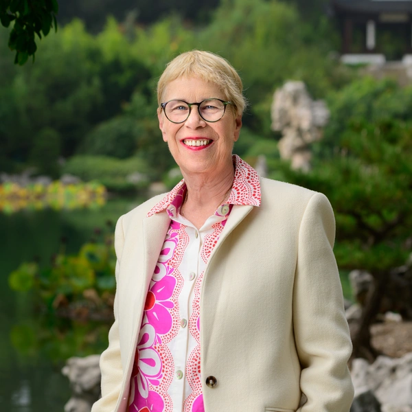 A smiling person in a pink floral shirt stands near water in a green garden. 