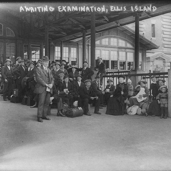 A black-and-white photograph of a large group of people under the awning of a building with large windows. At the top of the photo, in white writing, it reads: “AWAITING EXAMINATION, ELLIS ISLAND.”