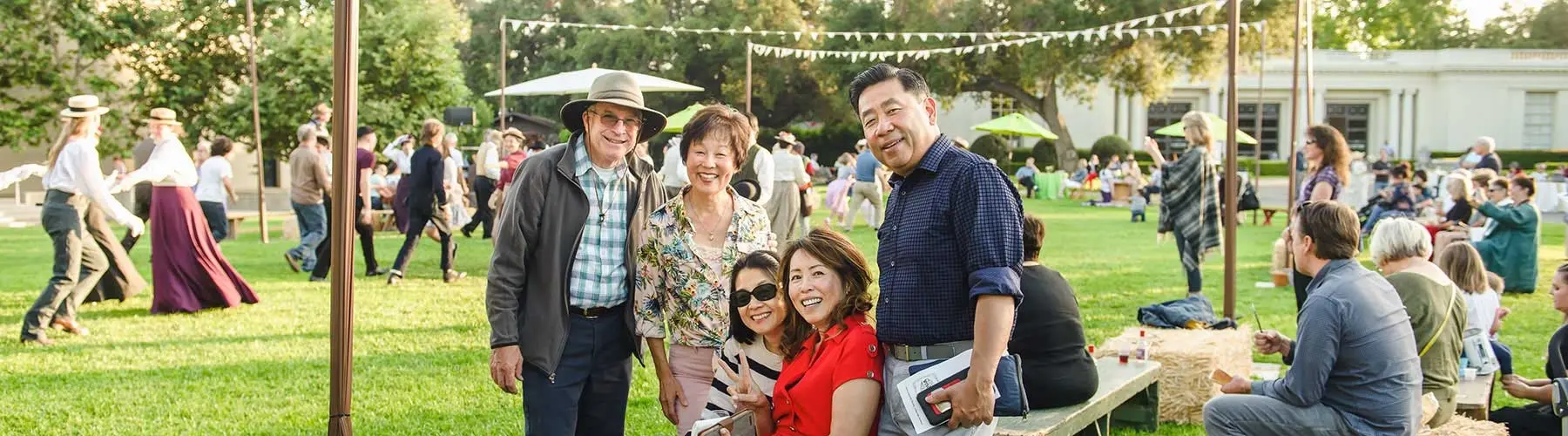 A group of five people pose for a photo at a daytime picnic on a large grass field. In the background are scores of people walking, dancing, or watching the action.