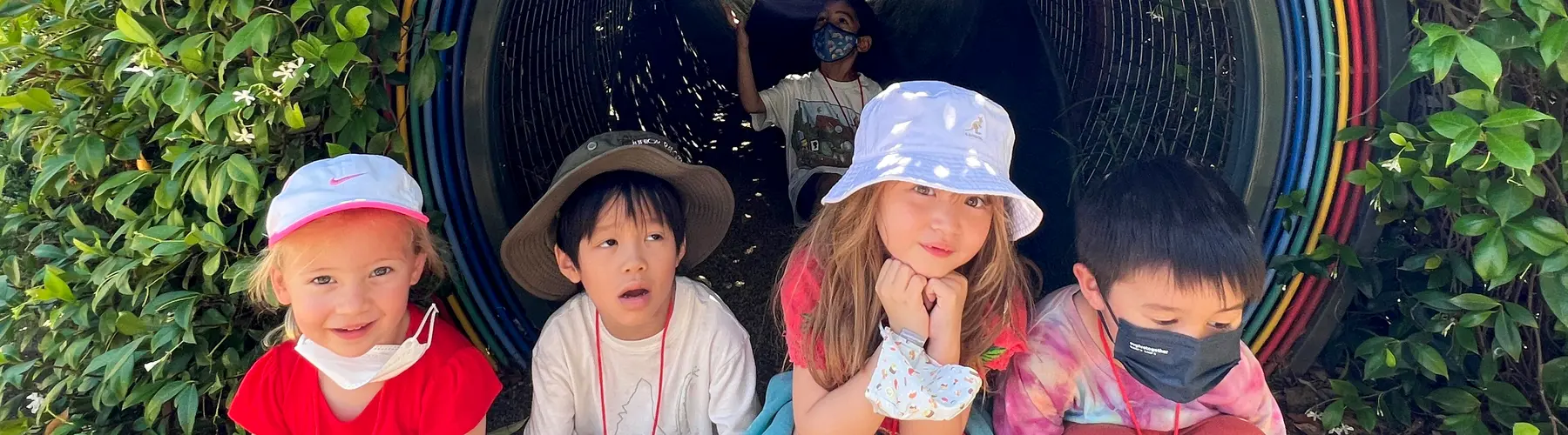 Children sit in front of a tunnel surrounded by greenery.	