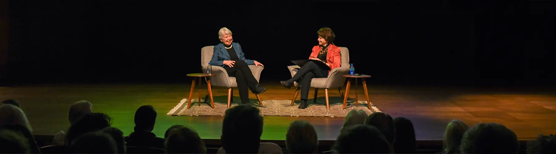 Two people sit in chairs on a stage in front of an audience.