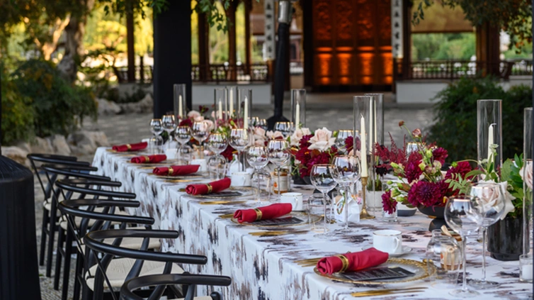 A dining table with place settings of red and light pink set up in an outdoor courtyard.