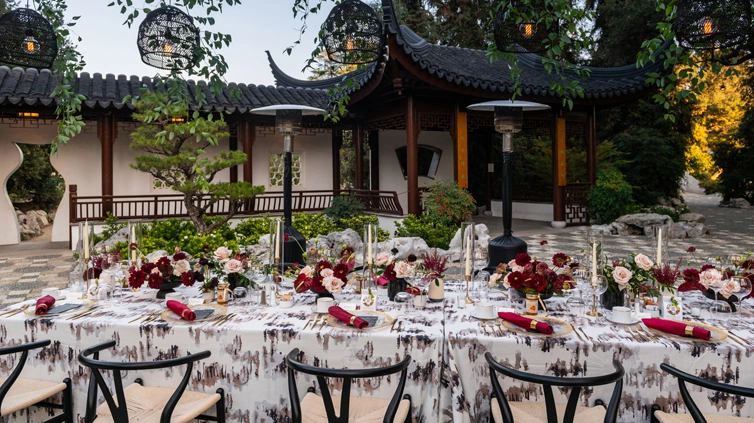 A dining table with place settings of red and light pink set up in an outdoor courtyard.