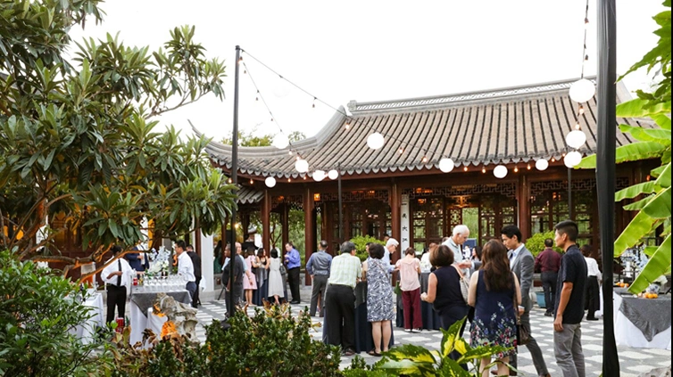 People are gathered in a courtyard for an outdoor event with string globe lights above.