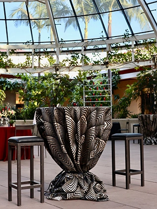 A round event table set up with a black and gold-patterned tablecloth under glass dome.