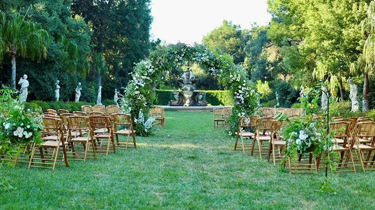 A plant-filled trellis separates rows of wooden chairs set up on a green lawn with a large garden fountain in the background.