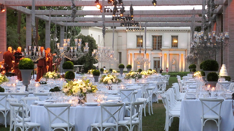 Attached to top of a wooden pergola are colored spotlights that shine down on white tables and chairs.