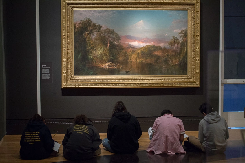 A group of people sit on the floor, in front of a painting of a landscape, in a dimly lit room.