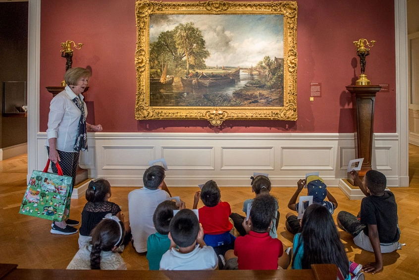 A group of young people sit on the floor, in front of a painting of a landscape, in a museum.