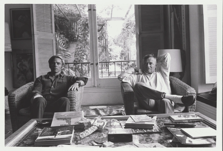 A black-and-white photo of two men in a living room, sitting behind a coffee table filled with books.