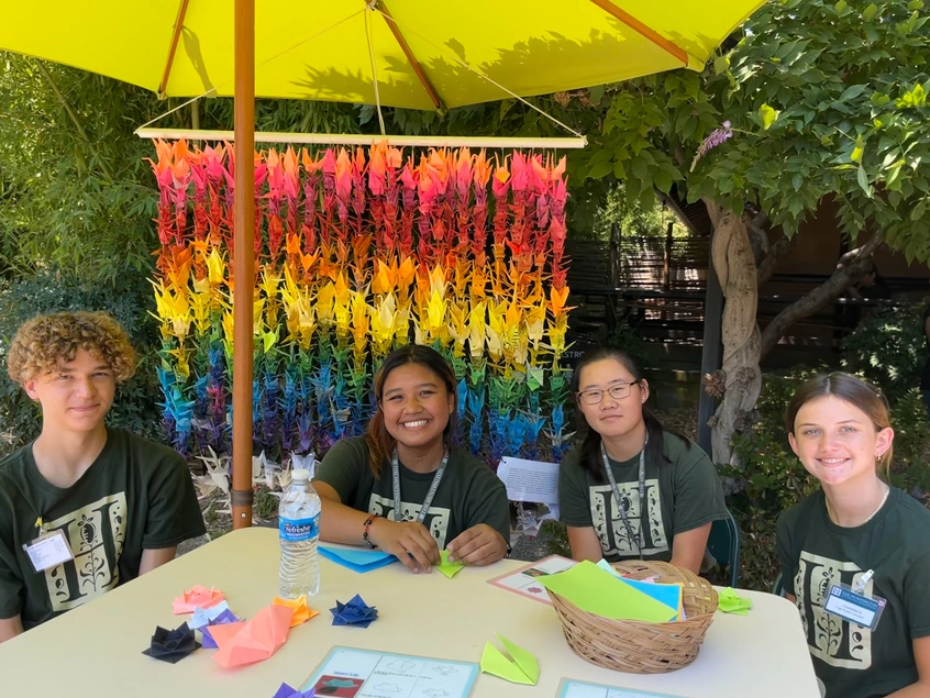 Four young people sit at a table with craft supplies, in front of a curtain of folded paper cranes.