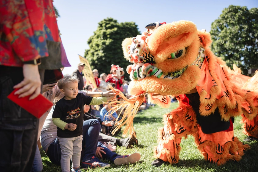 A child in a crowd interacts with a large lion puppet in a garden.