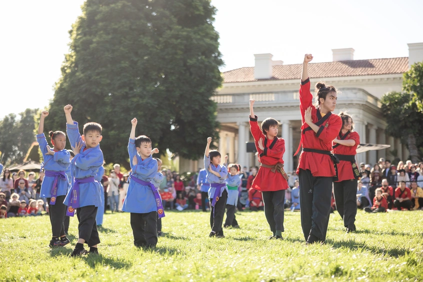 A group of children in blue outfits and adults in red outfits perform martial arts in front of a crowd on a grass lawn.