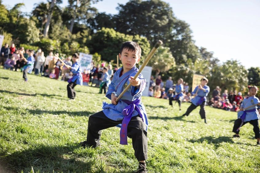 A group of children in blue outfits perform martial arts in front of a crowd on a grass lawn.