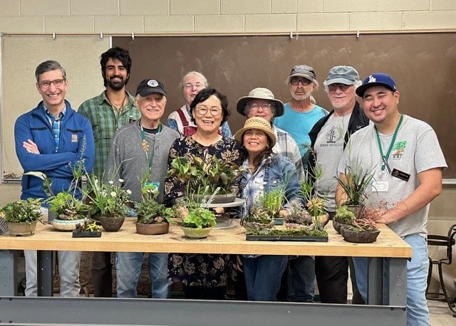 A group of people stand behind a table filled with bonsai.