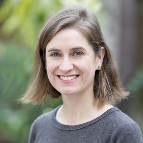 A smiling person with shoulder-length brown hair, wearing a gray sweater.