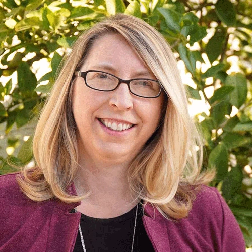 A smiling person with shoulder-length blond hair, wearing glasses and a magenta-colored jacket.