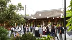 People are gathered in a courtyard for an outdoor event with string globe lights above.